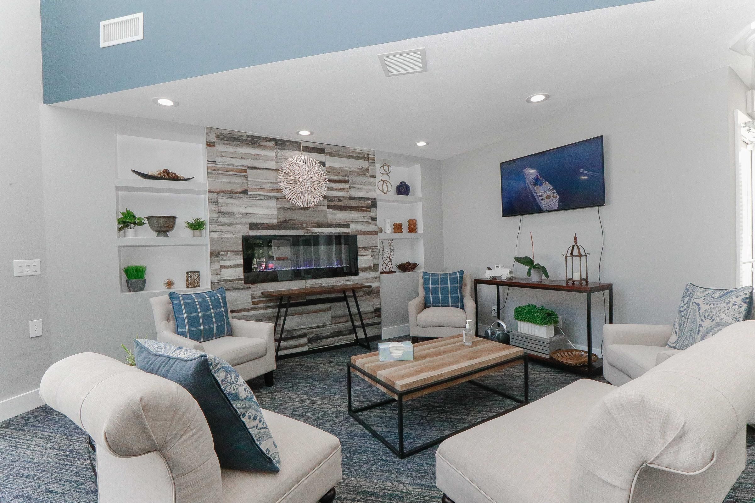 Modern living room with neutral chairs, blue accents, wall shelves, and a TV above an electric fireplace.