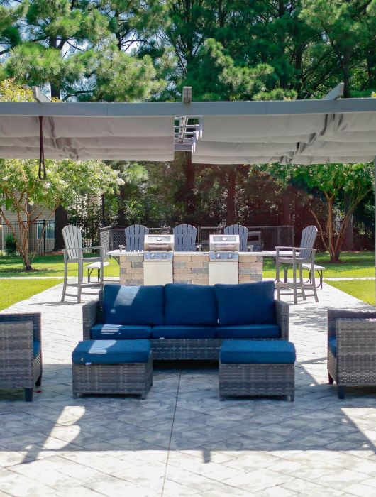 Outdoor patio with wicker seating under a pergola, grill stations, and tables set on a paved area surrounded by trees.