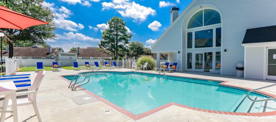Outdoor swimming pool beside a modern building with lounge chairs, tables, and a bright blue sky above.