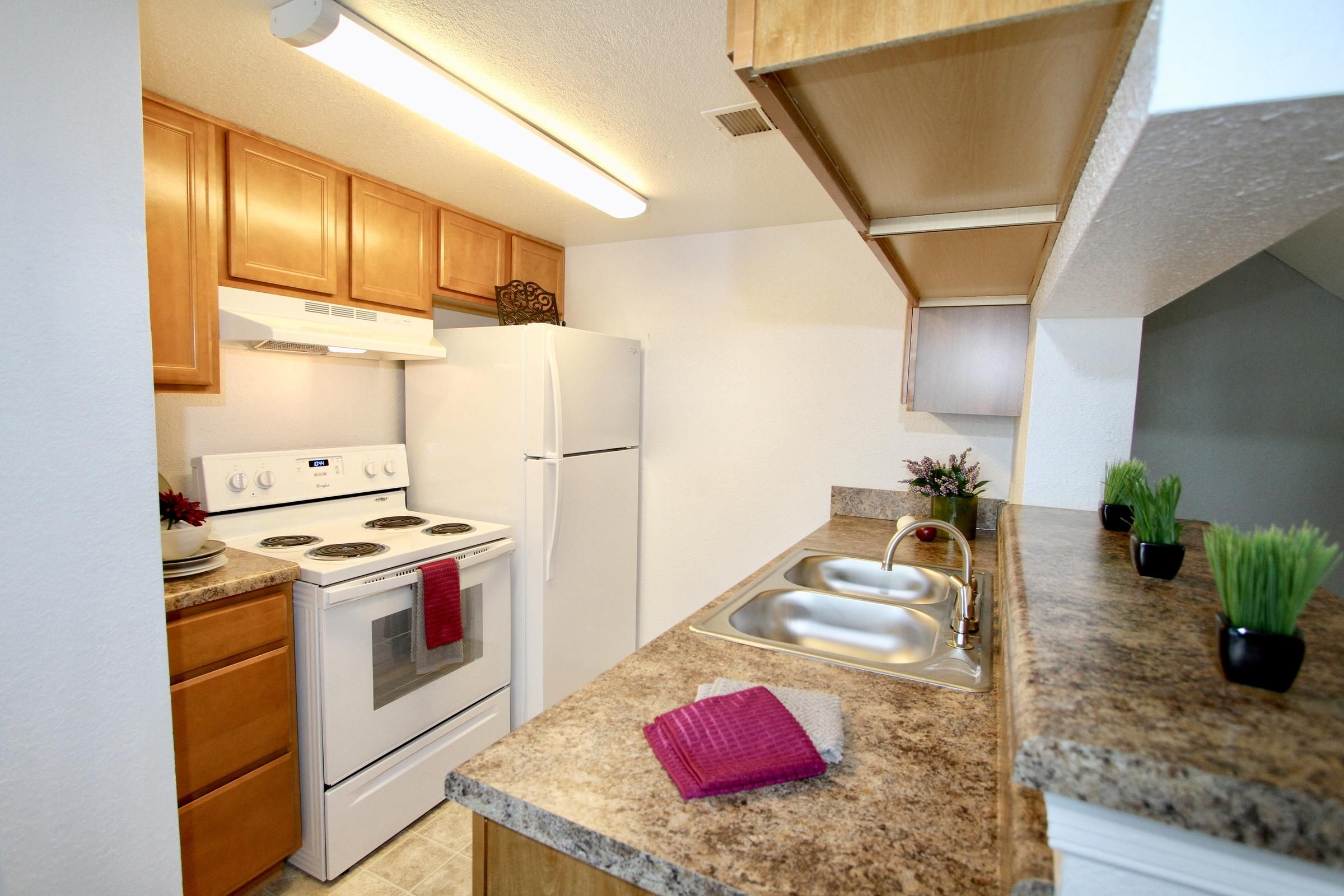 Small kitchen with wood cabinets, white appliances, double sink, and decorative towels and plants on the counter.