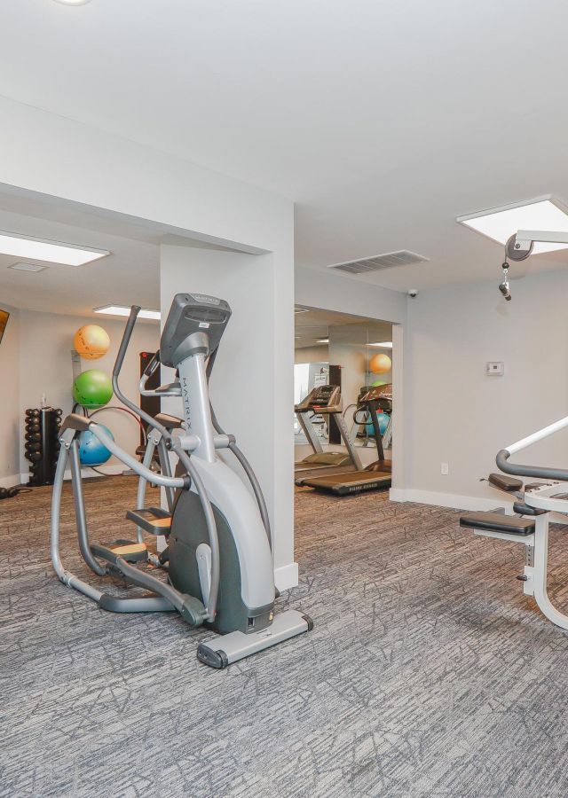 Small gym room with exercise machines, free weights, medicine balls, and bright overhead lighting.