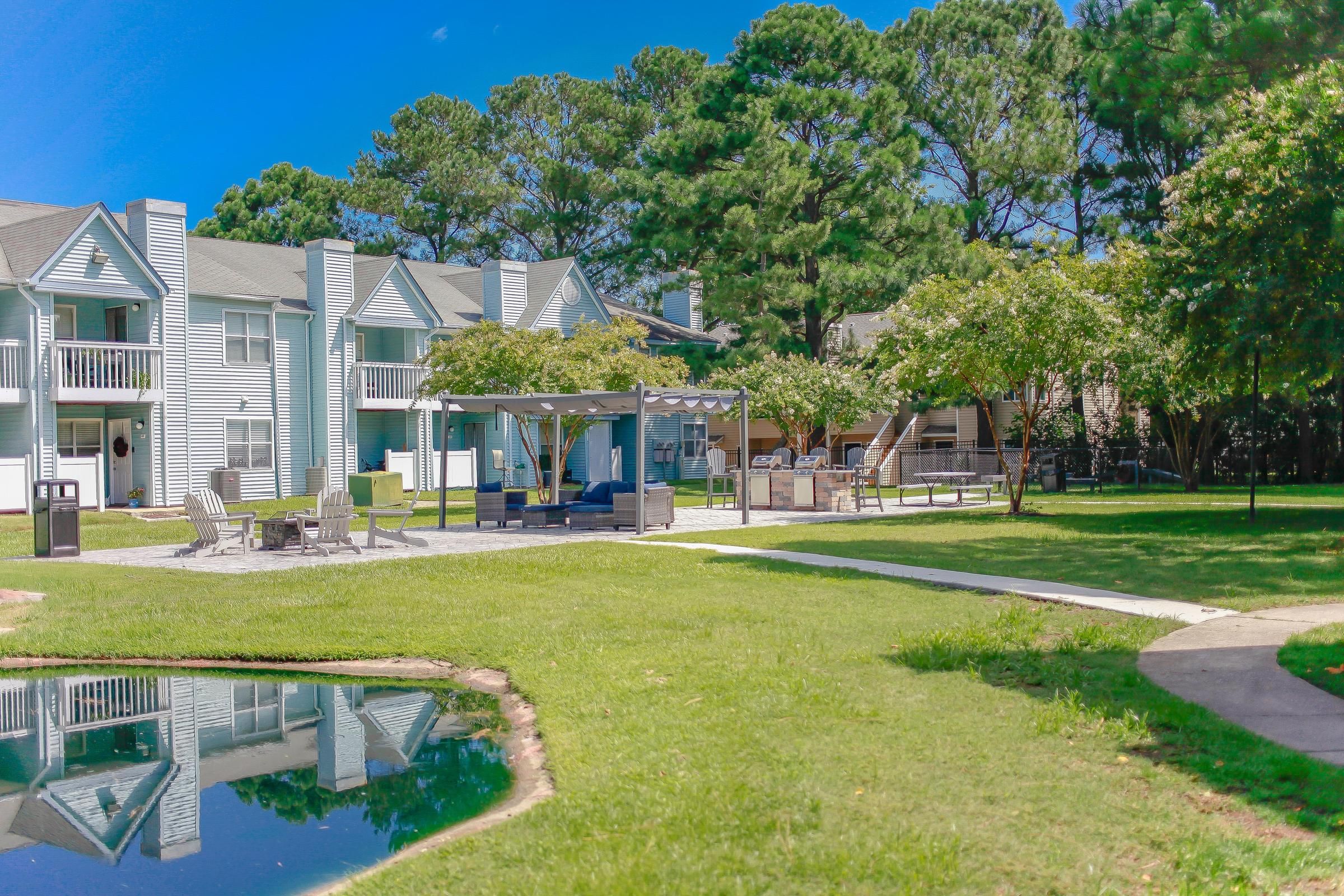 Apartment complex courtyard with patio seating, green lawn, trees, and a small pond under a blue sky.