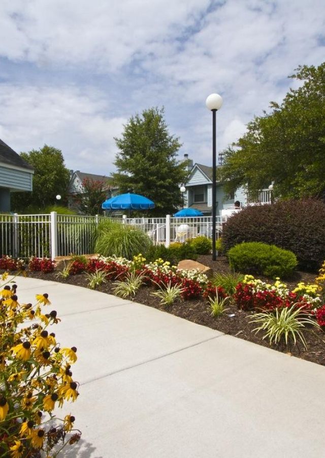 A sidewalk bordered by colorful flowers leads to a fenced area with blue umbrellas and trees under a partly cloudy sky.